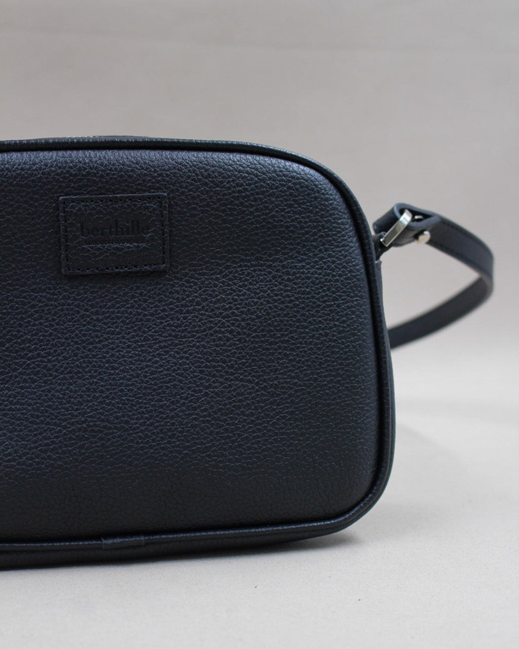 Gros plan logo gravé et attache bandoulière, minaudière cuir grainé noir Berthille