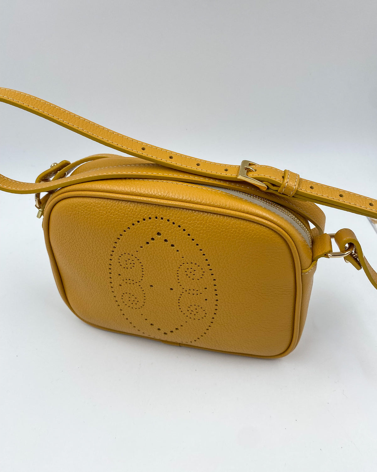 Pochette Trianon en cuir jaune moutarde vue de trois quarts, motif perforé bout fleuri et bandoulière réglable à boucle dorée, maroquinerie artisanale française