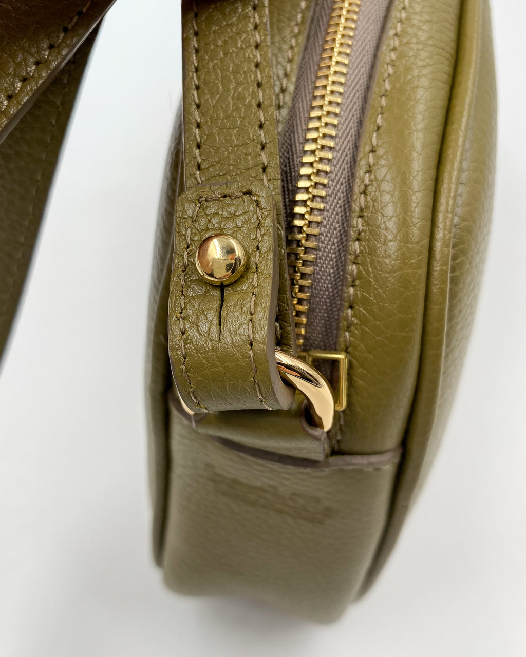 Détail de la pochette Trianon en cuir kaki : attache de bandoulière avec bouton et anneau dorés, coutures ton sur ton, fabrication artisanale française