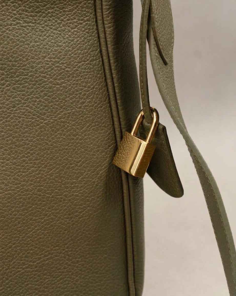 Détail du sac Trianon grand format en cuir kaki grainé : cadenas doré et clochette en cuir suspendus à la bandoulière, fabrication artisanale française