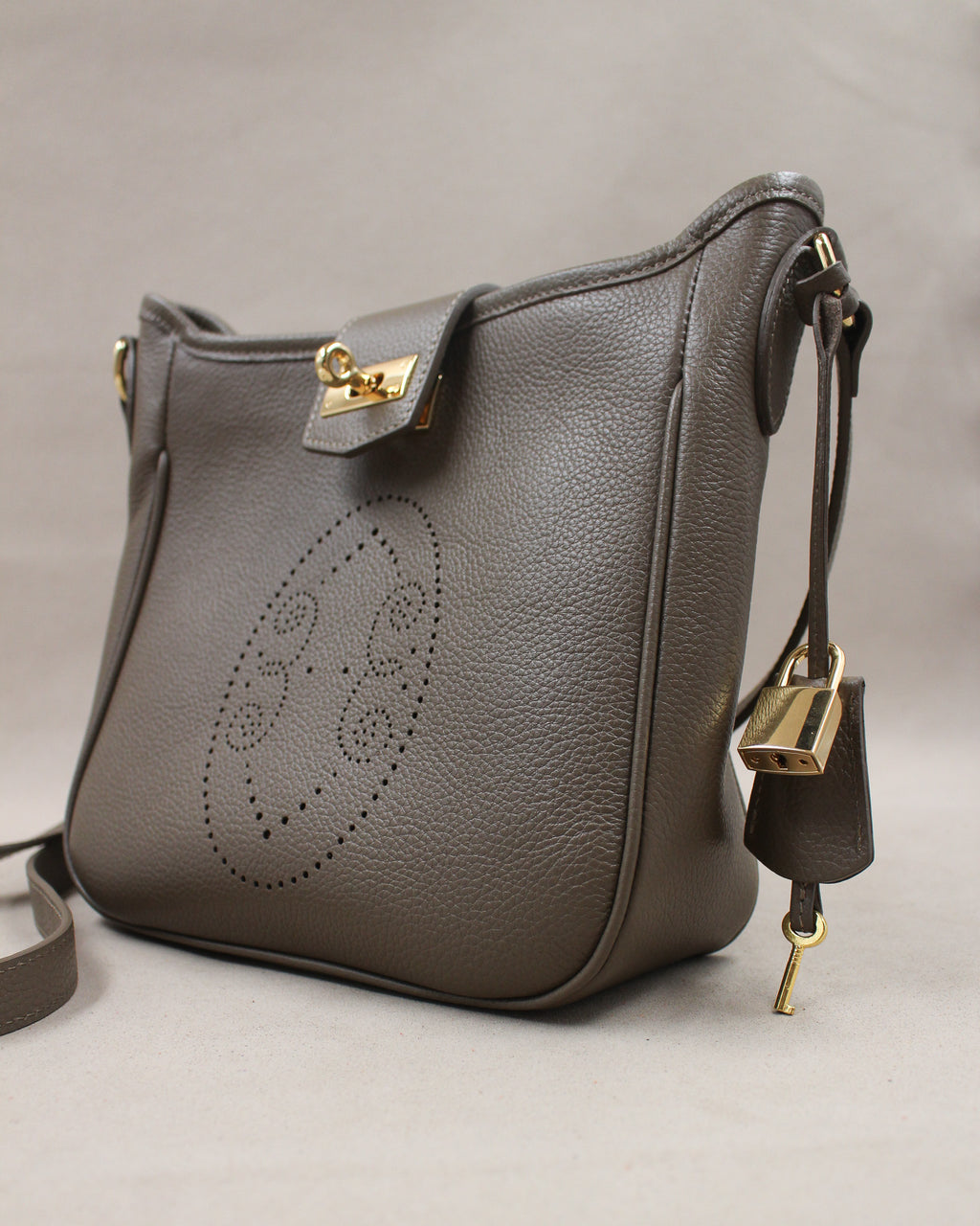 Sac Trianon en cuir gris vue de trois quarts, motif perforé bout fleuri, fermoir tournant doré, cadenas, clé et clochette en cuir, maroquinerie artisanale française