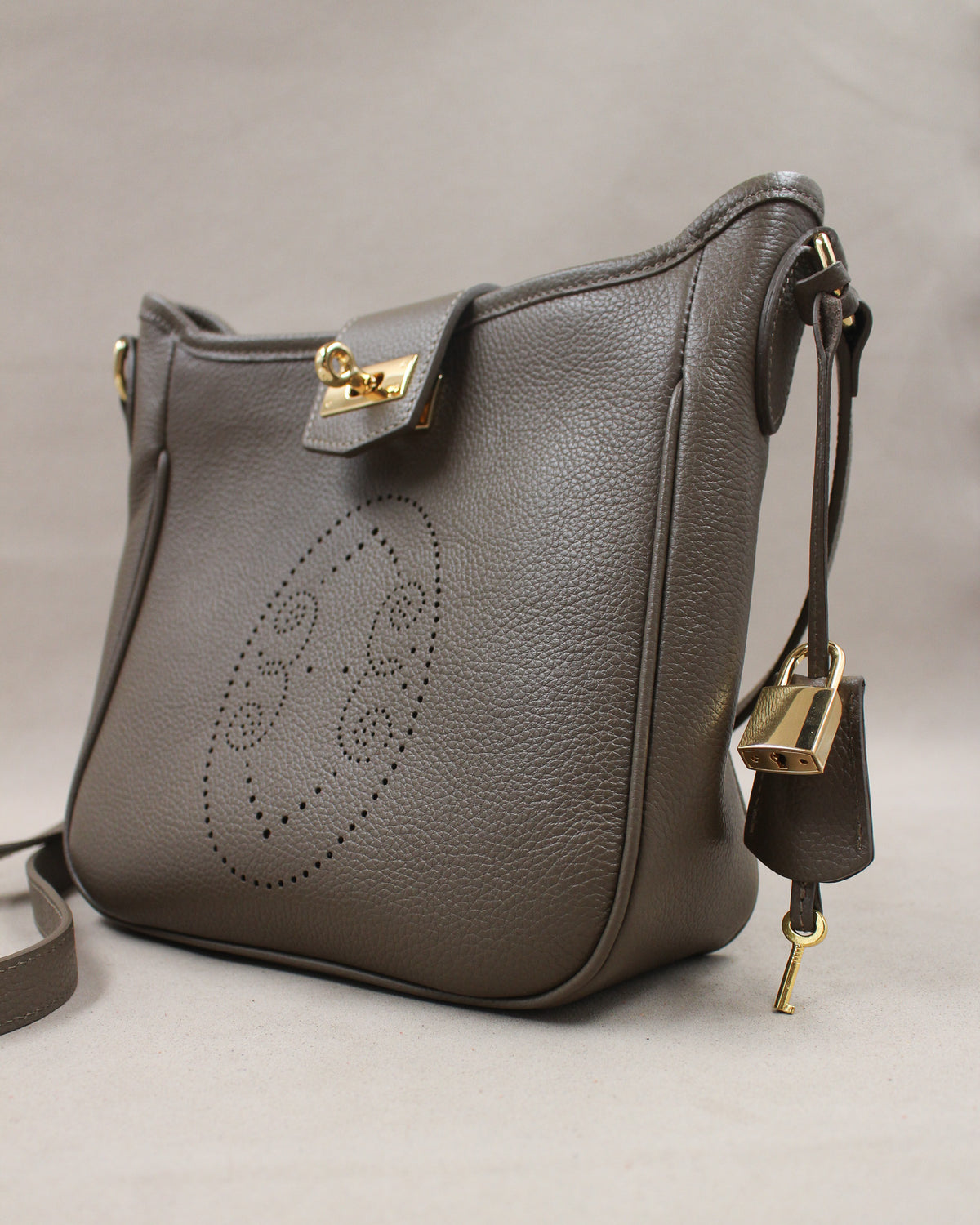 Sac Trianon en cuir gris vue de trois quarts, motif perforé bout fleuri, fermoir tournant doré, cadenas, clé et clochette en cuir, maroquinerie artisanale française