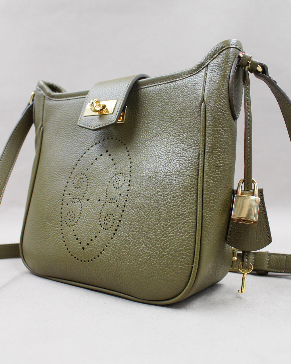 Sac Trianon en cuir kaki vue de trois quarts, motif perforé bout fleuri, fermoir tournant doré, cadenas, clé et clochette en cuir, maroquinerie artisanale française