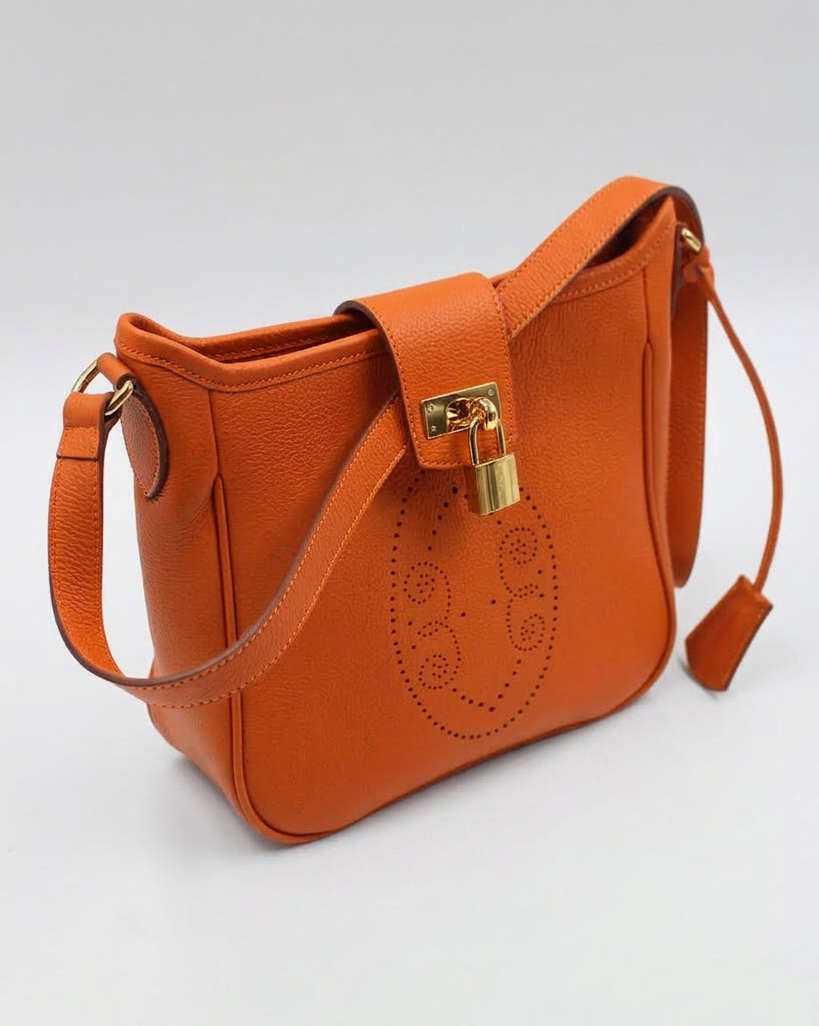 Sac Trianon en cuir orange vue de trois quarts, motif perforé bout fleuri, cadenas doré et clochette en cuir, maroquinerie artisanale française